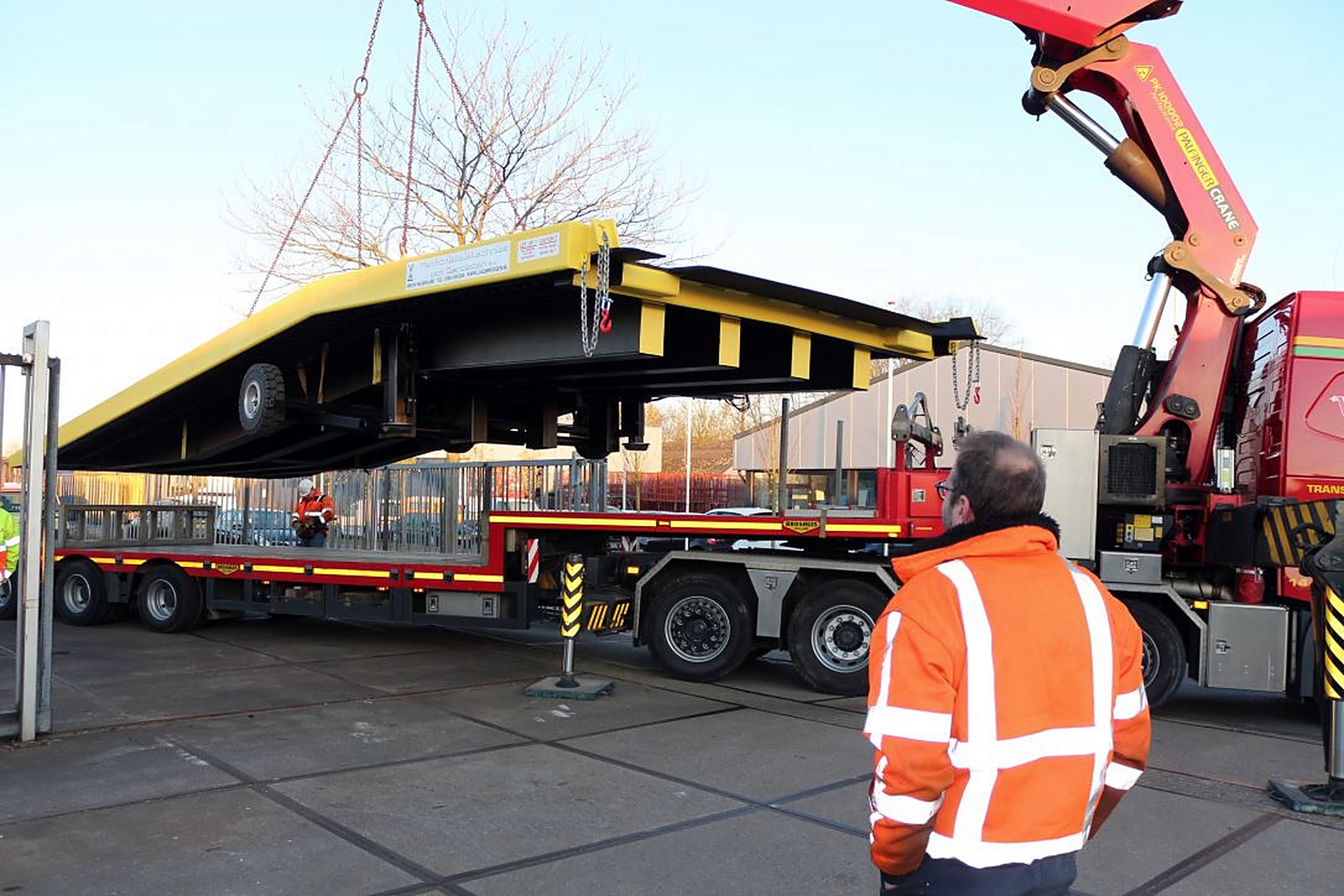 Een gele METEC laadbrug wordt met een kraan van een dieplader getild, een medewerker in oranje werkjas kijkt toe